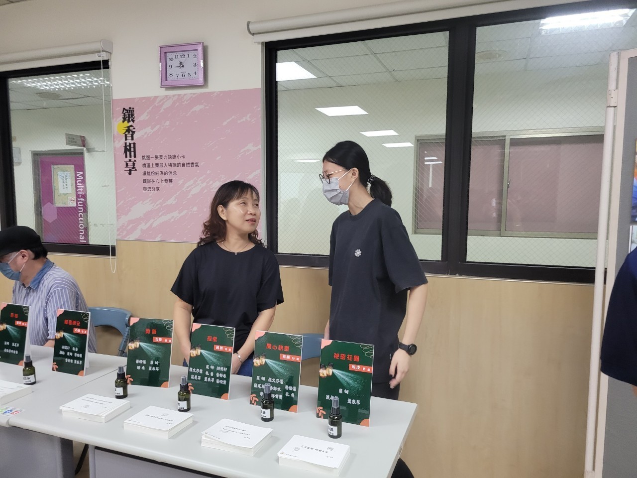 一位女生在一張桌子前與另一位戴口罩的女生交談