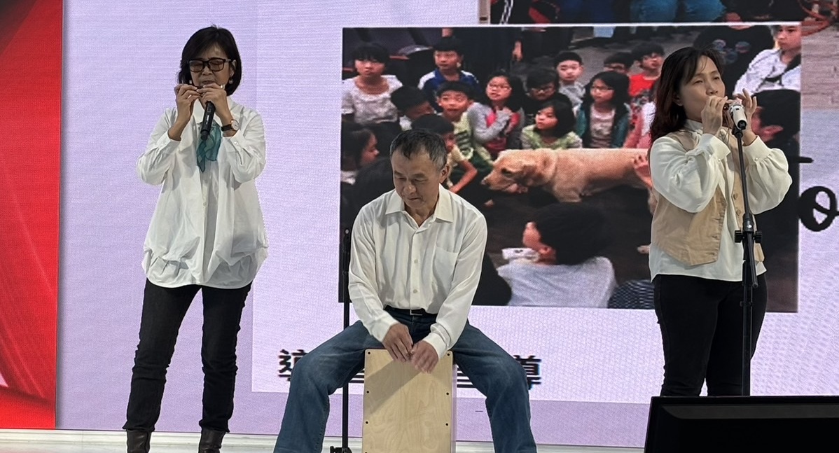 畫面中有三個人中間的男生正拍打著木箱鼓，在他左右兩邊的女生正陶醉的吹著口琴