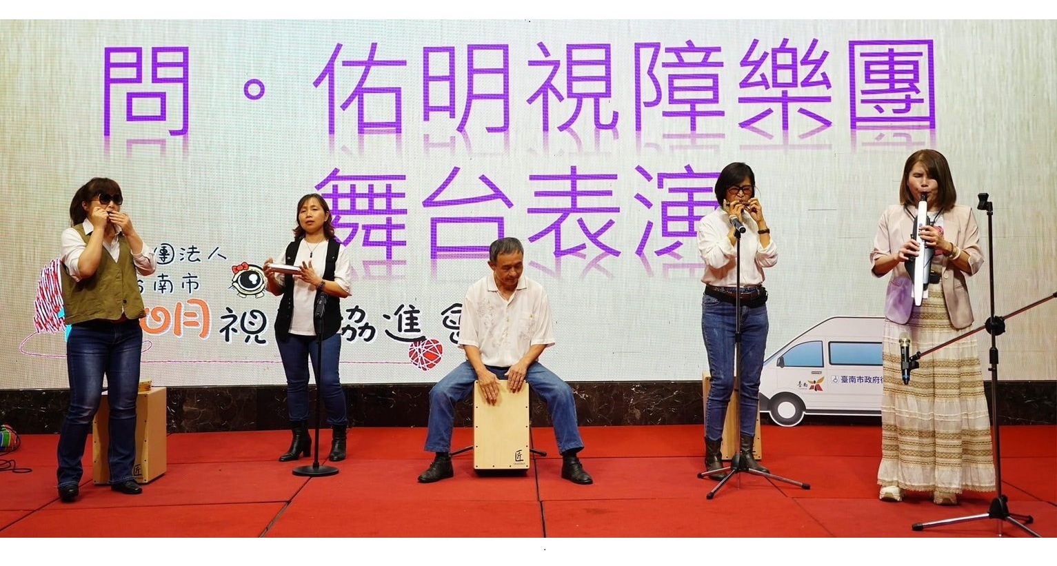 畫面中有五個人在紅色的舞台上表演，其中有兩支口琴、一顆木箱鼓、一支薩克斯風、還有一個沙鈴